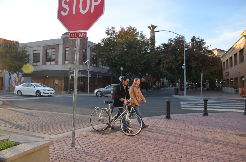 Photo shows a street corner with a stop sign and two adults walking side by side, one of the adults is walking with a bicycle