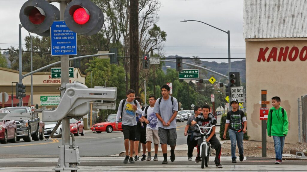 Photo shows a busy street with drivers of cars waiting at an intersection, and a group of children crossing at an intersection by walking and riding bikes